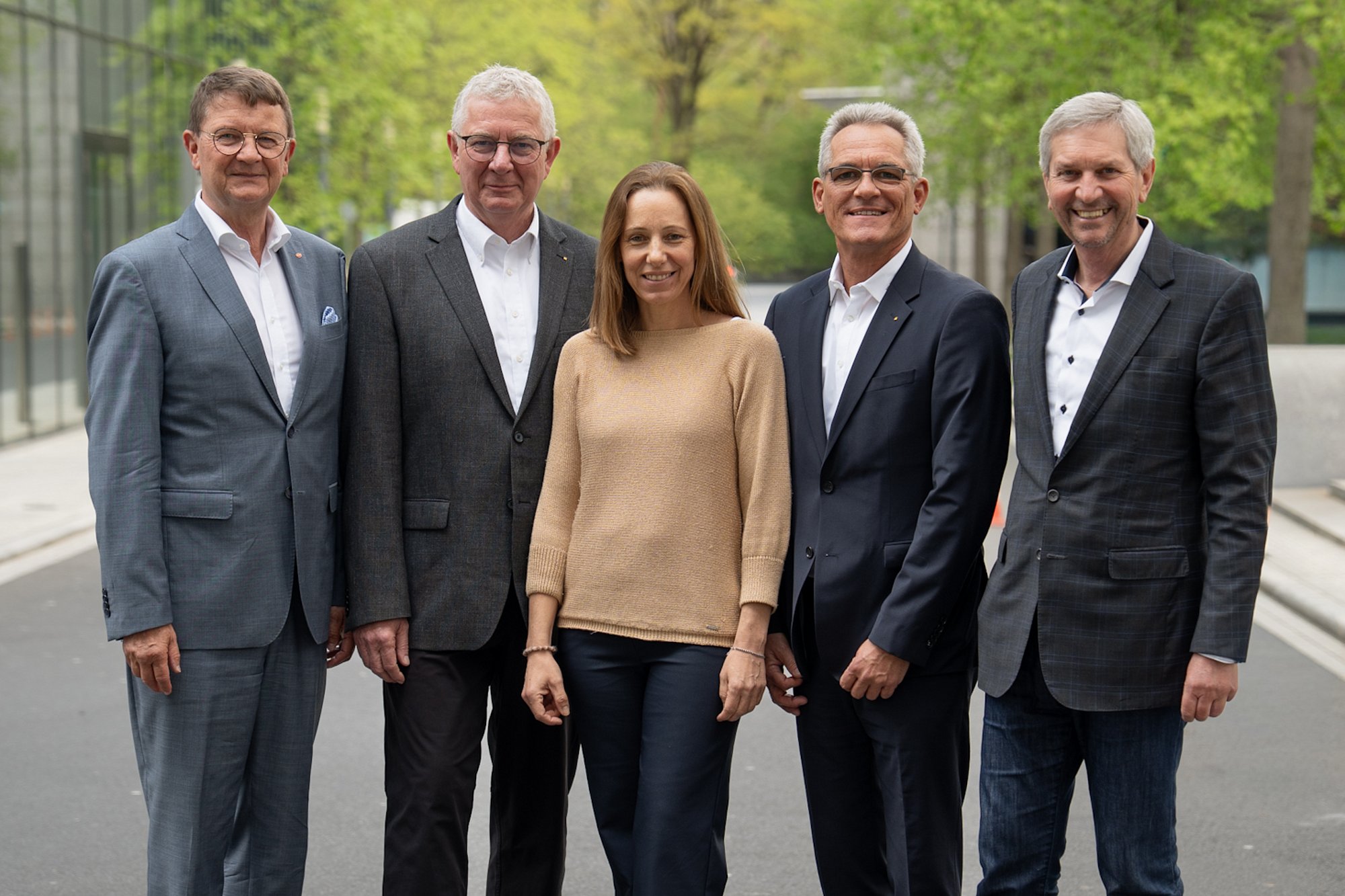 Fünf Personen in Geschäftskleidung stehen im Freien auf einem gepflasterten Weg zusammen und lächeln in die Kamera.
