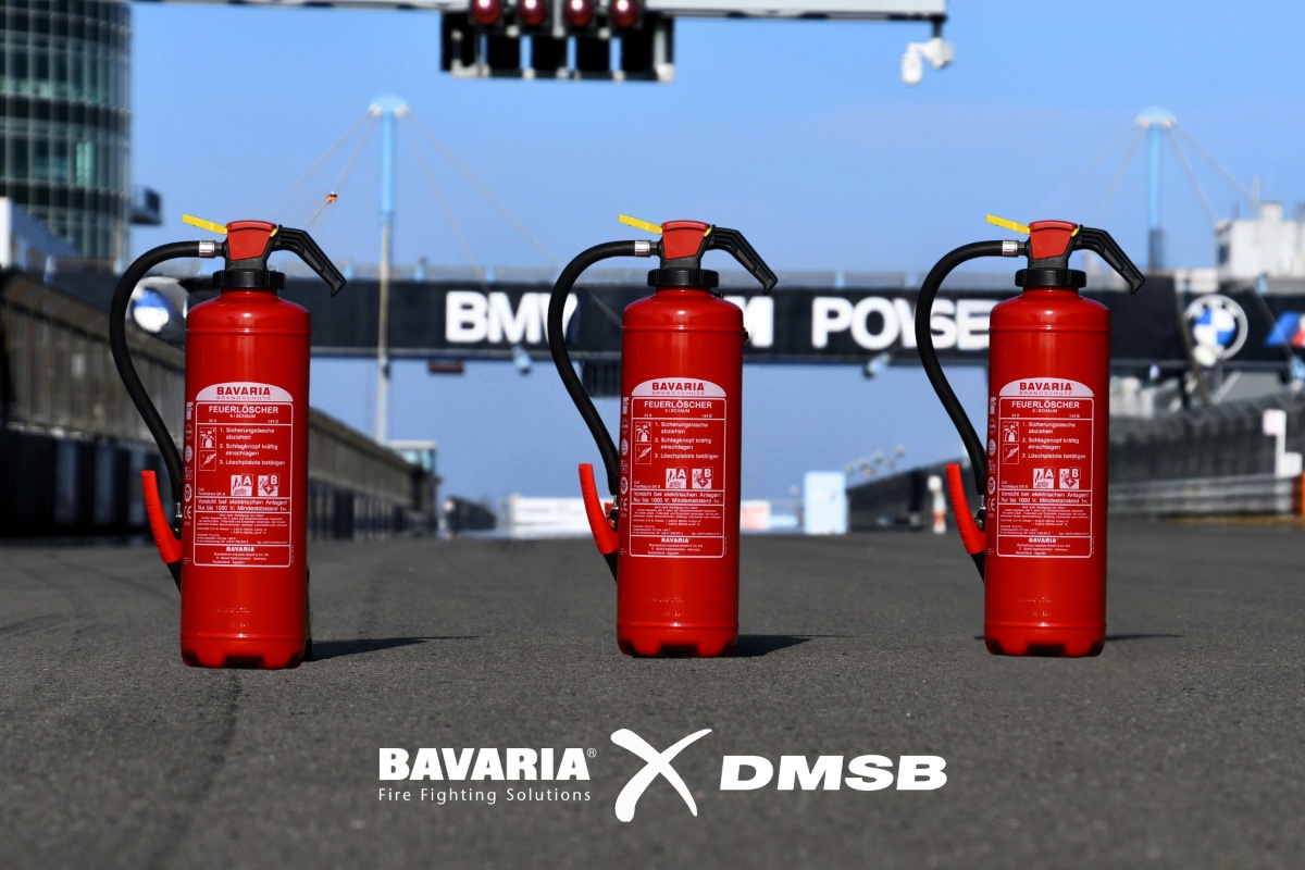 BAVARIA Feuerlöscher auf einer Rennstrecke x DMSB 3 rote BAVARIA Fire Fighting Solutions Feuerlöscher stehen auf einer asphaltierten Rennstrecke bei blauem Himmel. Unten die Logos von BAVARIA x DMSB in weiß.