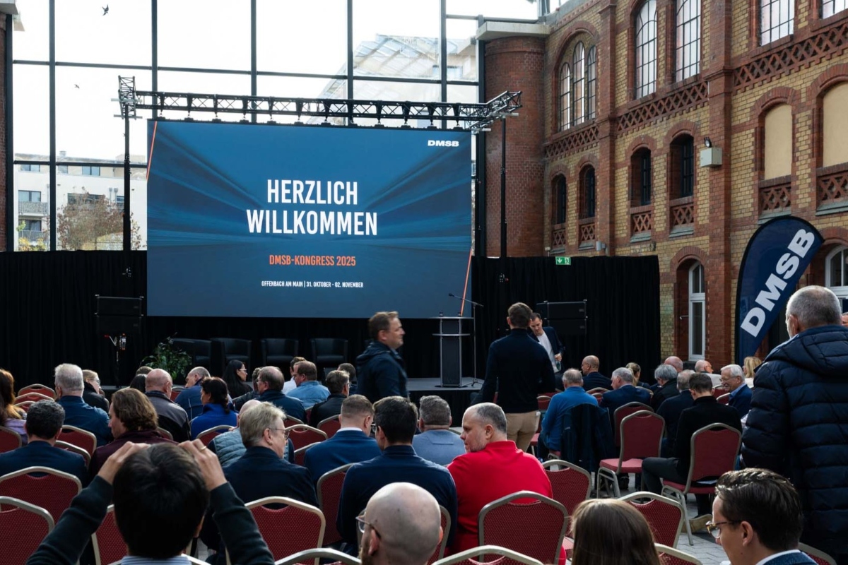 DMSB-Kongress 2025, Eröffnung Podium mit Rednerpult und Leinwand, davor Menschen in Sitzreihen, im Hintergrund Glasscheiben mit Sicht nach draußen. Auf der Leinwand steht: Herzlich Willkommen. DMSB-Kongress 2025. Offenbach am Main | 31. Oktober - 02. November 2025.